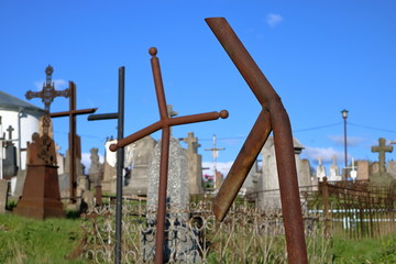 Stary cmentarz katolicki z pordzewiałymi, pokrzywionymi krzyżami, niektóre bez jednego ramienia, w tle kamienne stare nagrobki, błękitne niebo, słonecznie - obrazy, fototapety, plakaty