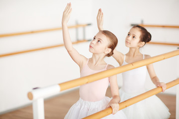 Small group of little girls in ballet dresses looking at their raised hands while training in...