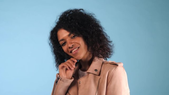 Portrait of woman smiling to camera, flirting, winds hair on finger and seductively bites her lip. Beautiful young mixed race trendy girl with afro hairstyle. 4k