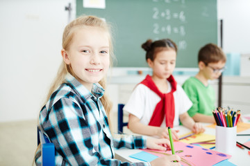 Fototapeta premium Happy blond schoolgirl smiling at you while sitting by desk and drawing at lesson
