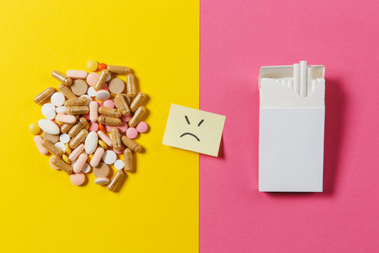 Medication White Colorful Round Tablets Arranged Abstract White Pack Cigarettes On Yellow Color Background. Paper Sticker Sheet Sad Smile Face. Treatment Choice Healthy Lifestyle Concept. Copy Space.