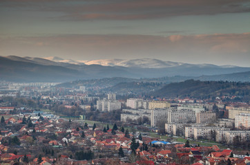 Elevated view of single-family houses and grey Eastern European socialist housing estates in Miskolc with wooded hills and snow-capped sunlit Bukk range of North Hungarian Mountains at winter Hungary