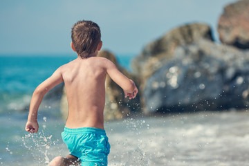 Cute Caucasian boy is running in the water along the sea shore against big boulders.