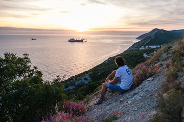 Fototapeta premium Unrecognizable men sit on hill and admires the sunset on mountain with seascape view Back view.