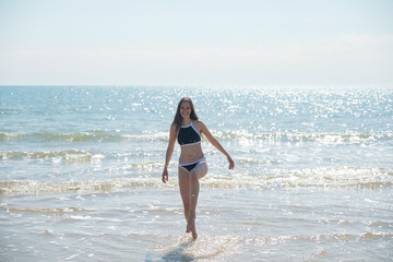 Fototapeta premium Brunette woman with long hair in blue bikini walks along the sea on a sunny day and smiles.