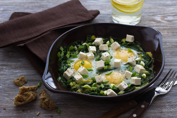 Green shakshuka with spinach, leek and feta in a ceramic frying pan on a wooden table with bread, selective focus. Delicious healthy home food