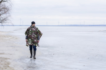 Young man walking on beach watching the frozen water in a military rain coat