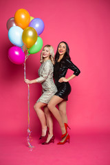 Obraz na płótnie Canvas Funny women. Two happy and cheerful beautiful girl friends in beautiful dresses posing and having fun with helium balloons in hands on a pink background.