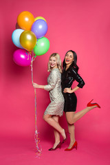 Obraz na płótnie Canvas Funny women. Two happy and cheerful beautiful girl friends in beautiful dresses posing and having fun with helium balloons in hands on a pink background.