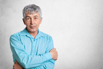 Indoor shot of confident mature man in formal shirt, keeps hands crossed, looks directly into...