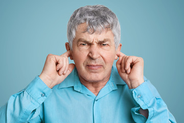Portrait of upset annoyed mature man plugs ears with fingers, dressed in formal shirt, poses against blue background, doesn`t want to hear anything. Irritated elederly man ignores someone