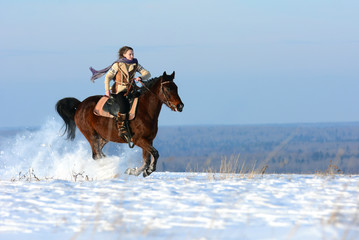 Winter horse riding on snowy field, covered dry snow with beautiful view from snowy peaks. Horseback riding with snow flying around under horse legs.  Happy girl on powerful horse running gallop.