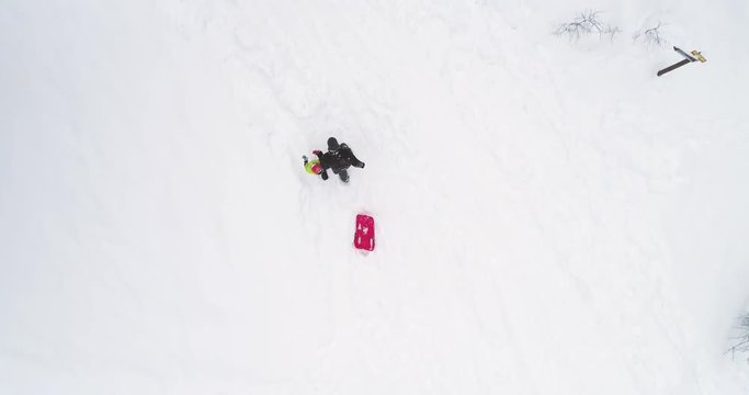 Winter Overhead aerial top view over father pulling red bobsled on snowy rise with child.Dad, son or daughter, bobsleigh on snow.Family people have fun together outdoors.4k straight-down perspective