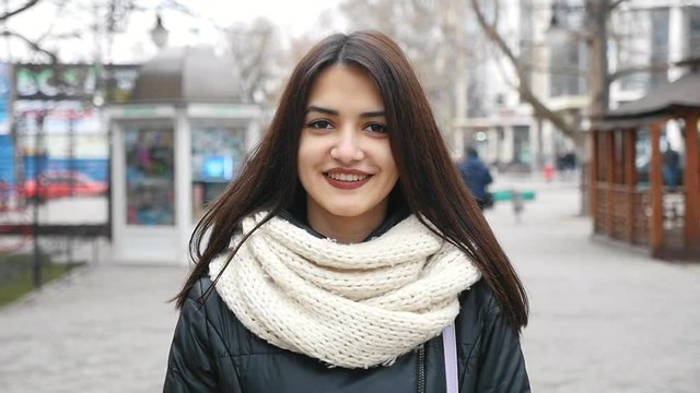   An optimistic view of a gorgeous girl with deep eyes who smiles happily in a street in winter in slow motion. She wears a big white scarf.