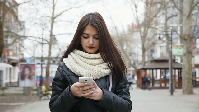 A splendid view of an elegant young girl with long loose hair who strolls in a street and looks at her mobile in winter in slow motion.