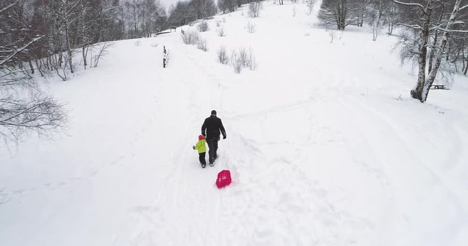 Winter aerial  follow father pulling red bobsled on snowy rise with child.Dad, son or daughter, bobsleigh on snow.Family people have fun together outdoors.4k drone flight video