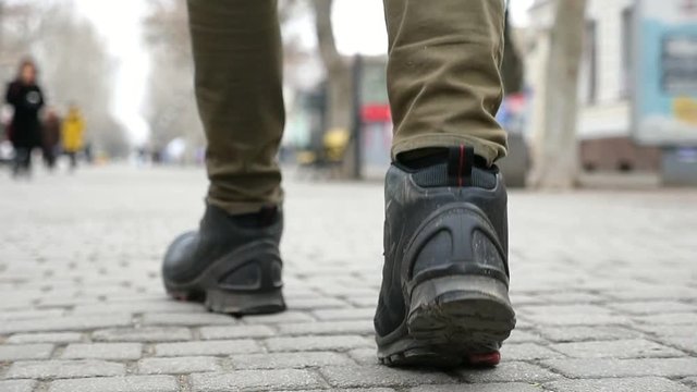 An original view of a young man`s feet walking on a tiled city street in winter in slow motion. The feet are in trendy black boots.  
