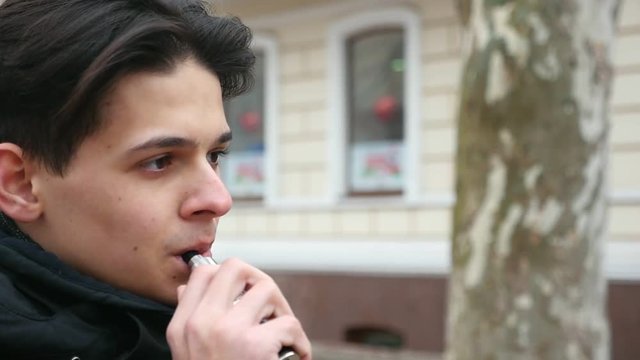 A stylish young man with a trendy haircut stands by a house and smokes an electronic cigarette in winter in slow motion. He feels relaxed and happy