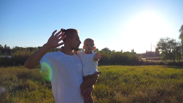 Portrait of happy family waving their arms into camera at the meadow. Young dad holding his son on hands at nature. Father and child spending time together. Nature landscape at background. Close up