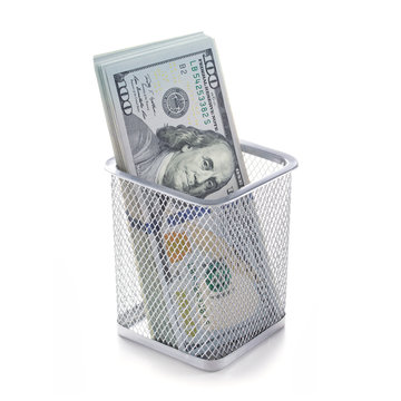 Money in a basket on a white isolated background. Garbage can full of dollars tipped on its side with money spilling out.