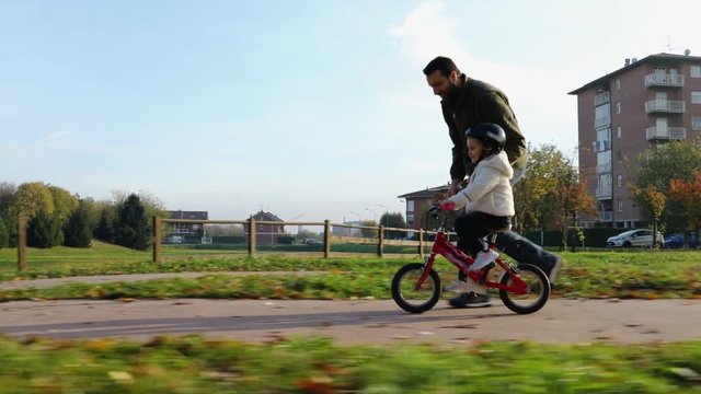 father teaching daughter to ride bike at urban park. Child girl learning biking with the dad's help.Family and childhood concept.Sunny autumn day.Side gimbal follow 4k video 