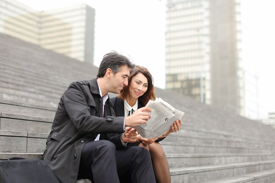 Engineers Male And Female Find Article About Company In Newspaper, Colleagues Sitting On Stairs Reading. Americans Wearing Business Clothes Smiling Communicating. Concept Of Fashionable Cl