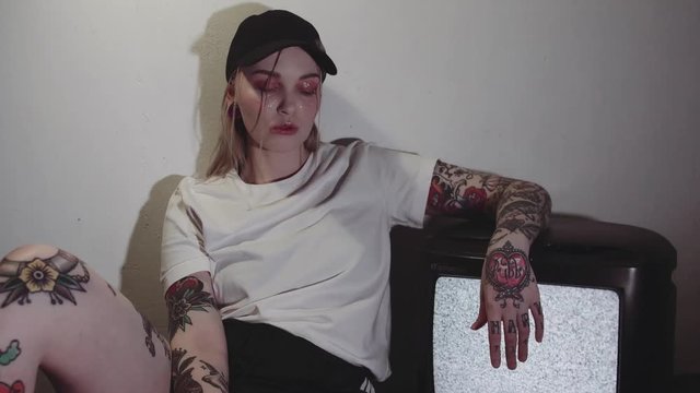 Punk tattooed caucasian woman with glitter on face in white shirt and black cap sitting near working static tv set in dark room