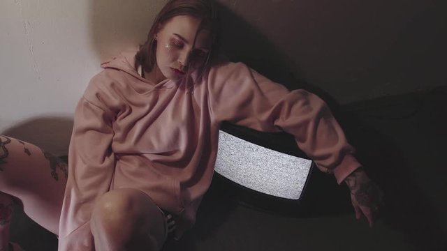 Hipster tattooed caucasian girl with glitter on face in pink hoodie and black cap sitting near working static tv set in dark room