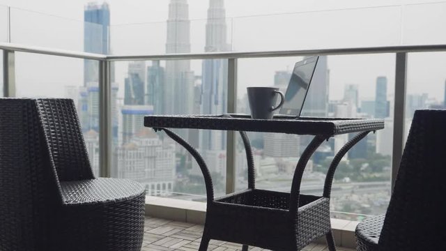 Teo chairs and a table with cup of coffee and open laptop at terrace with beautiful view over the city. Luxury apartment balcony and interior.