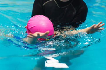 A little girl of European appearance in a pink rubber cap learning to swim in the pool