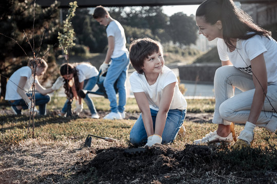 Young environmentalist. Cheerful nice positive woman looking at the boy and smiling while helping him to plant a tree