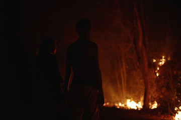 Back view of young tourists couple silhouette standing hand by hand together in darkness watching local farmers burning forest on purpose. Couldn't do anything with disaster. Human relations concept.
