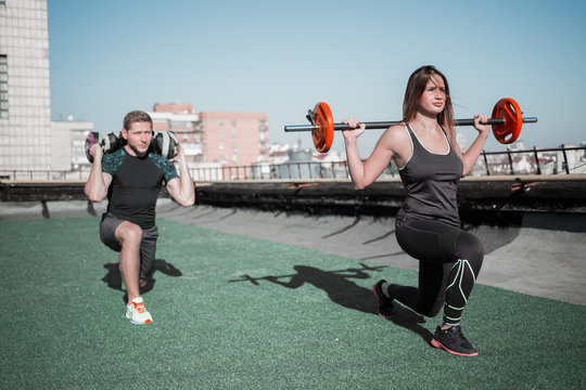 Young people doing workout on the roof. Sportive lifestyle man and woman doing lunges with barbell.