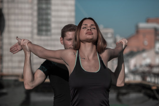 Man and woman working out outdoors. Fitness on the roof concept, man helping young female to stretch arms and shoulders.