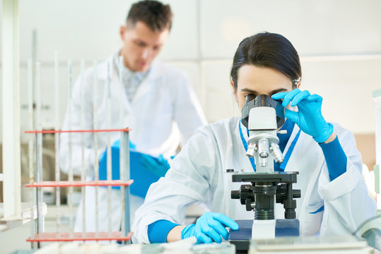Confident dark-haired scientist wearing white coat and rubber gloves sitting at desk and using microscope, her male colleague standing behind and taking notes