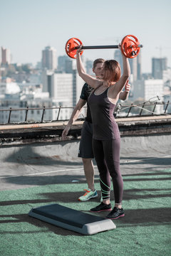 Young woman liting a barbell on the roof of the building. Sport exercises on the top of a building in the city.