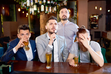 Nervous bearded friends gathered together in pub after work in order to watch football match and drink beer, one of them covering eyes with hand