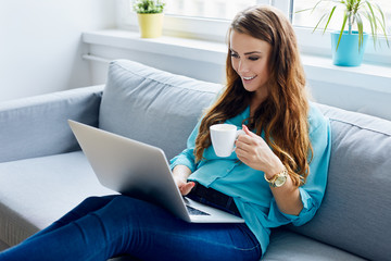 Attractive young woman sitting on sofa at home using laptop and holfing cup of coffee