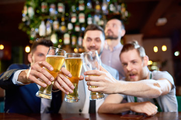 Group of cheerful friends clinking beer glasses together while sitting at bar counter and  celebrating end of work week, focus on foreground