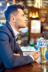 Profile view of handsome young man wearing stylish suit sitting at bar counter after hard working day, drinking beer and watching rugby match