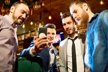 Low angle view of cheerful friends looking through funny photos on smartphone while spending evening in modern pub