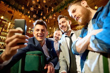 Smiling bearded friends taking selfie with help of smartphone while having long-awaited gathering...