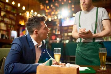 Profile view of young entrepreneur sitting at table and making order while spending evening in modern cafe, bearded red-haired waiter taking notes