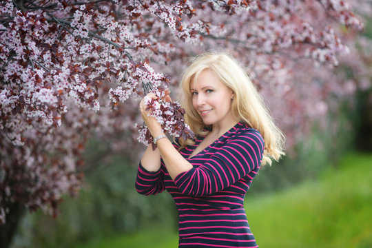 Happy smiling Caucasian blond woman with long hair in purple fedora hat near blossoming plum cherry tree, enjoys the blossom
