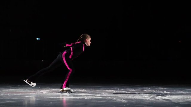 A little girl skater in a black sports suit and white skates performs a spinning stand in the arena of the ice stadium. The leg is directed back of the hand to the sides. Rotating the gyroscope