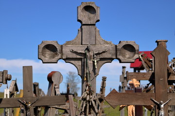 Drewniany krzyż z wizerunkiem ukrzyżowanego Chrystusa, w tle nieostrych wiele innych krzyży, drewnianych i metalowych, prostych, przekrzywionych, w plenerze, w tle nieostre drzewo, błękitne niebo © Wioletta