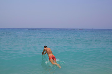 Young guy jumps into the blue water with sea foam. Turquoise water with white pebbles on the beach. Sea line meets the horizon making beautiful panoramic view. Handsome man dive in pink swim trunks.