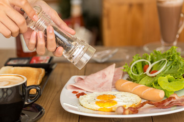 Hands of woman holding a Bottle of pepper on Morning American breakfast with sunny side up eggs, bacon, toast and coffee