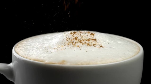 Pouring cinnamon powder into cappuccino coffee cup . Black background. Close-up shot. Slow motion
