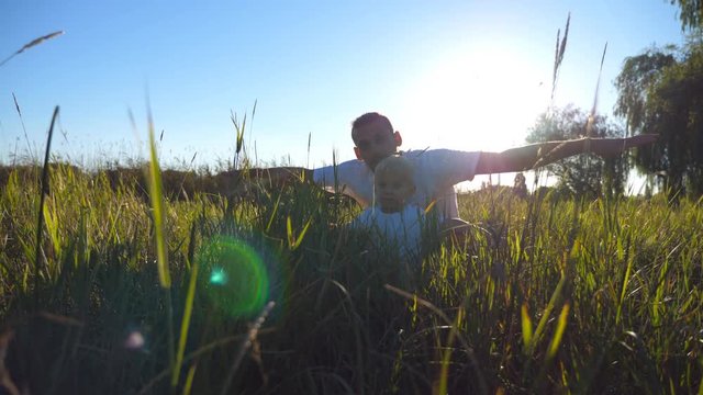 Portrait of young father and little son sitting on green grass and playing raised hands as airplane. Family spending time together and enjoying nature. Sunlight at background. Close up Low angle view